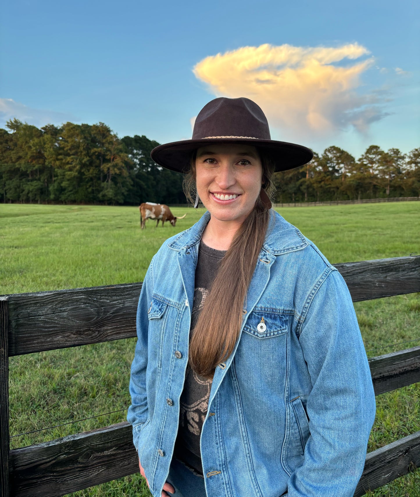 Woman in a denim jacket and wide-brimmed hat standing in a field with a cow and wooden fence.