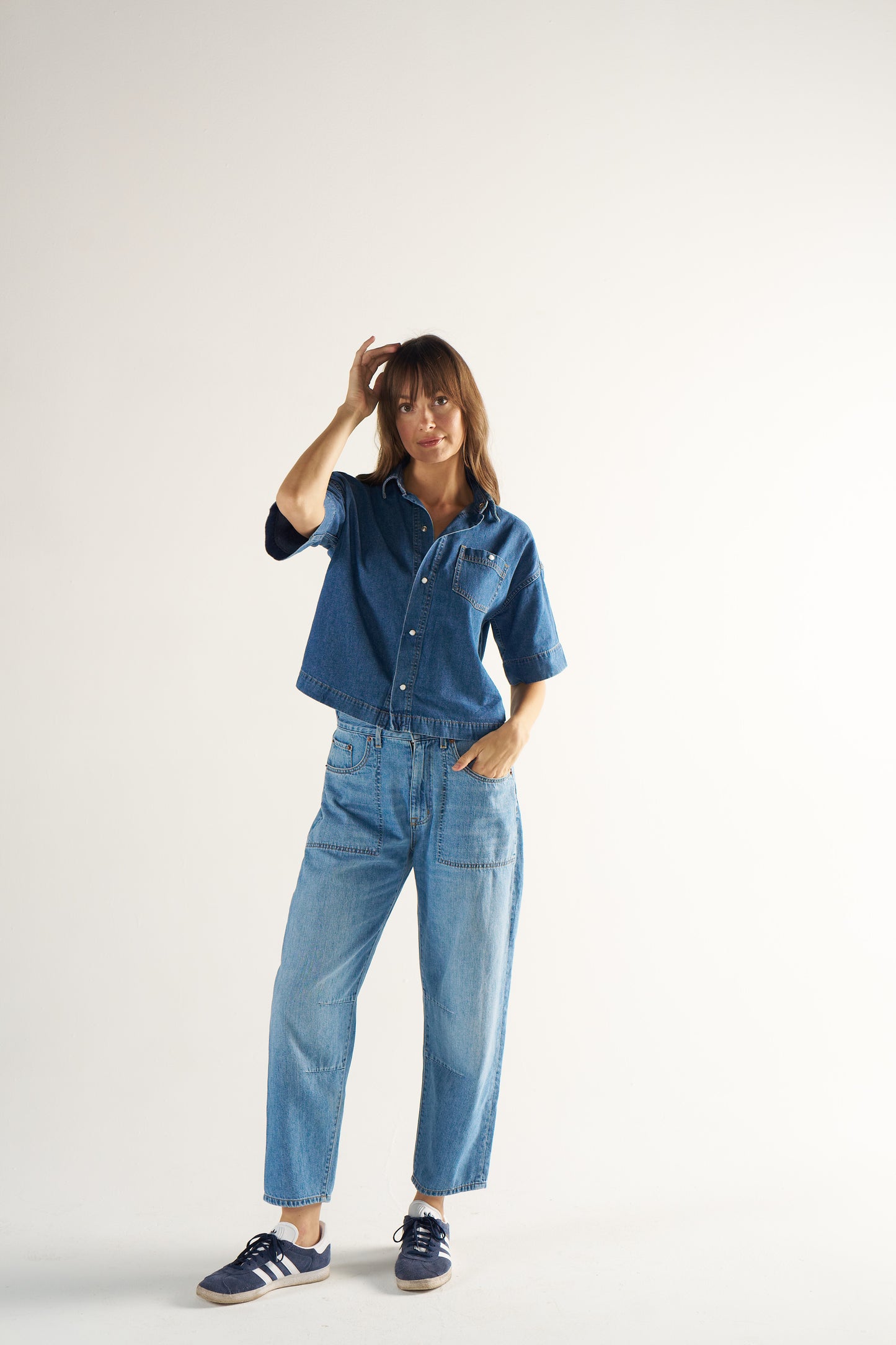 Woman wearing a blue denim shirt and jeans with navy Samba sneakers on a white background