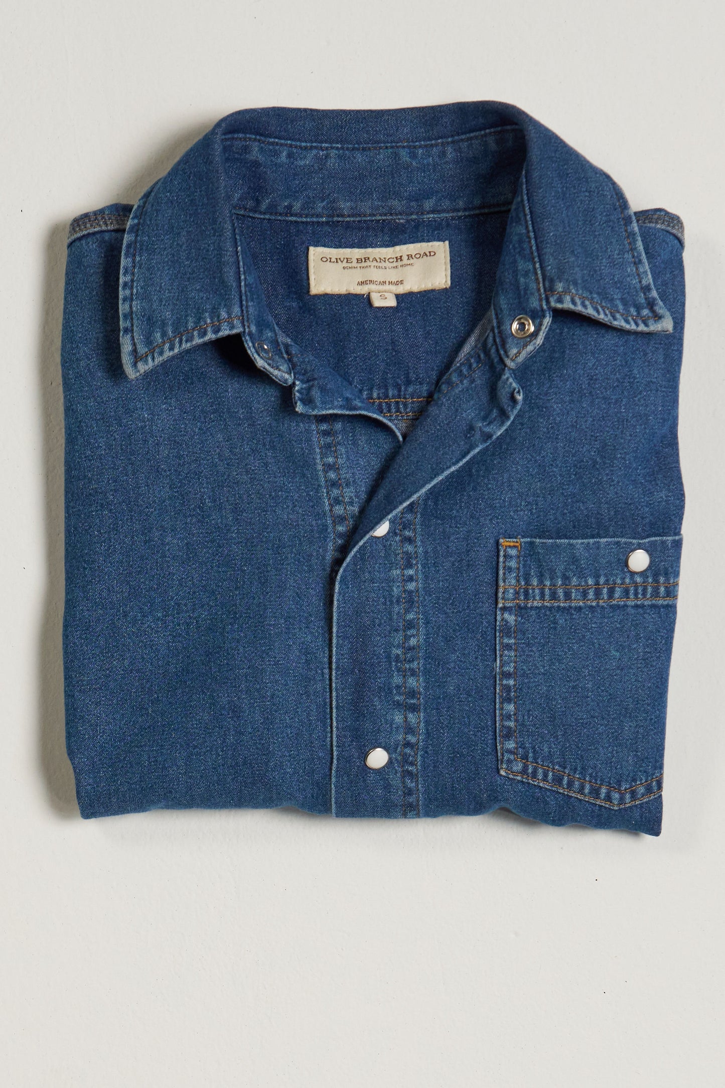 Folded indigo blue denim work shirt with pearl snaps on a light gray background