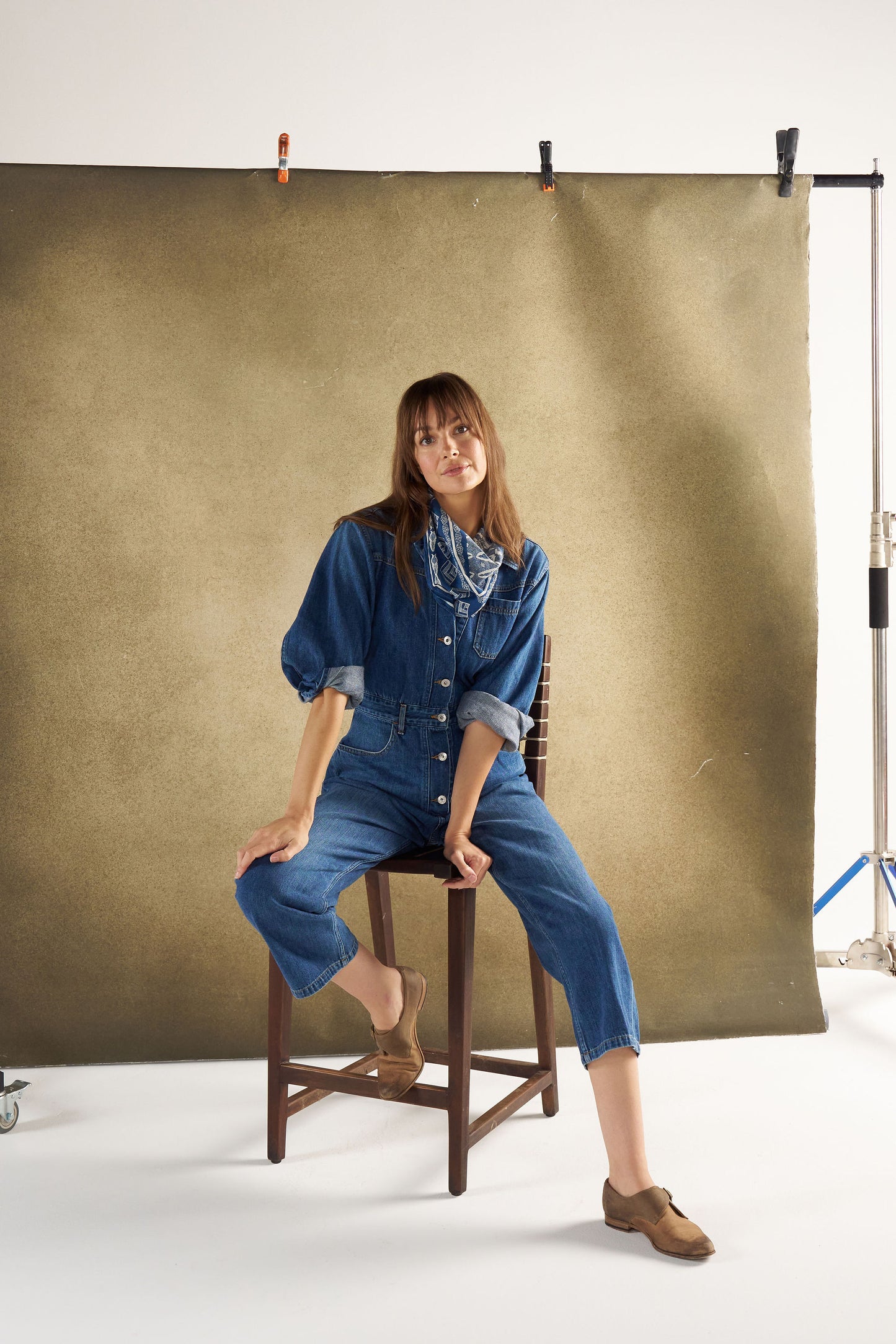 Woman in a blue denim outfit sitting on a chair against a brown backdrop