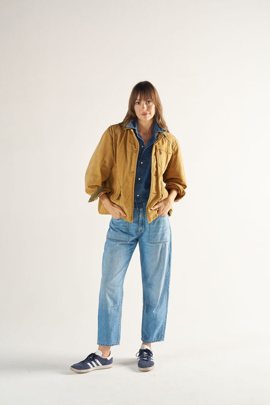 Woman wearing a mustard jacket, blue shirt, and jeans on a white background