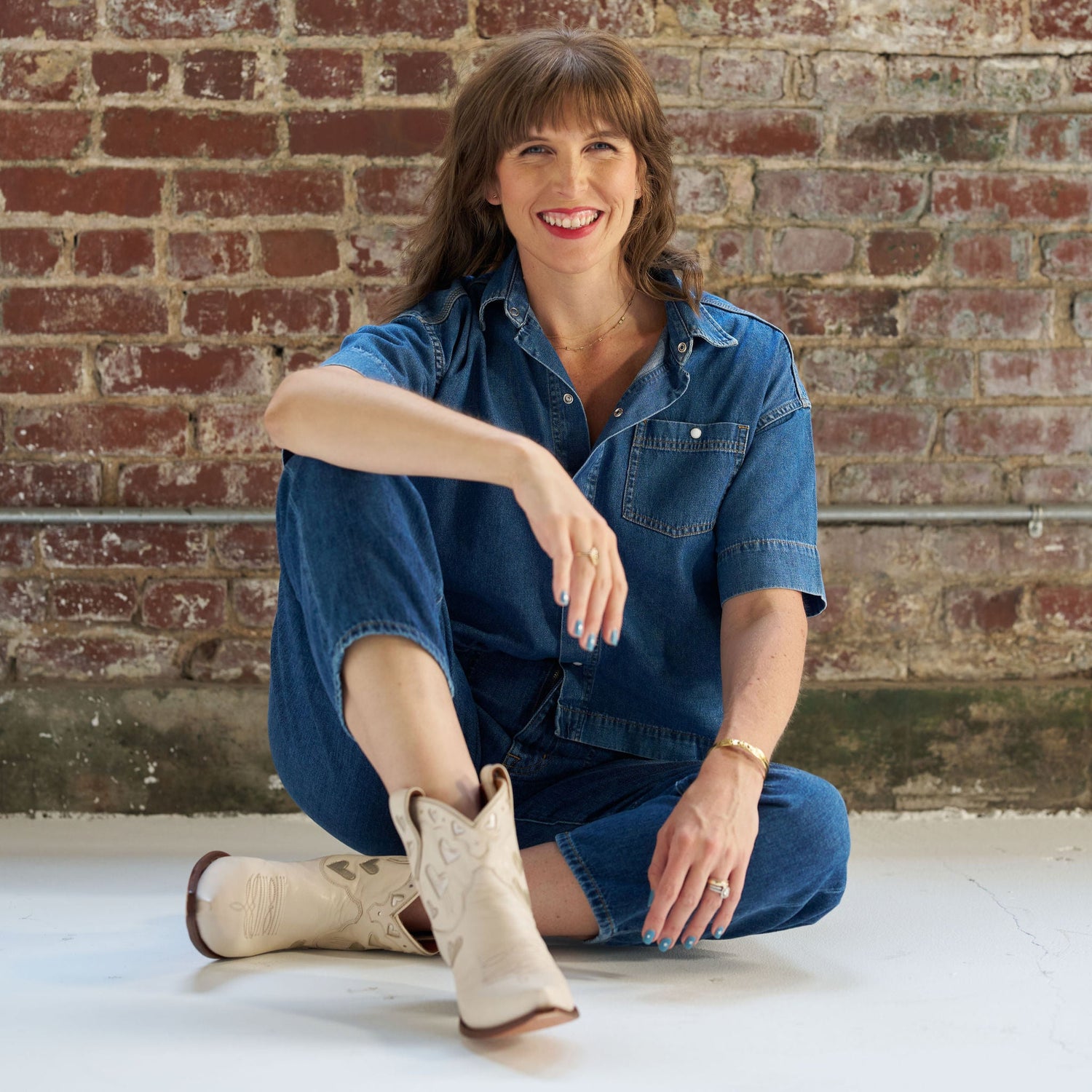Woman sitting on the floor against a brick wall wearing a denim outfit and white Miron Crosby boots.
